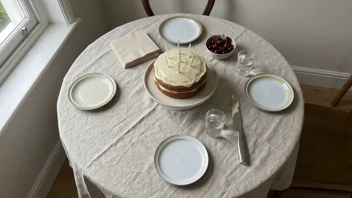 A round table by a window laid for a birthday, with a homemade cake and candles in the middle